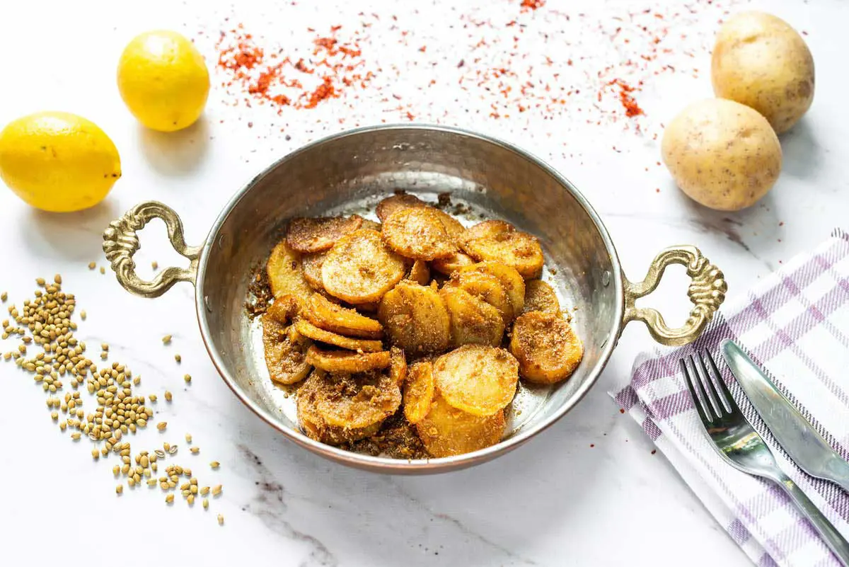 Gebratene Kartoffelscheiben in einer Kupferpfanne, umgeben von Zitronen, Koriandersamen und rotem Gewürz auf einem Tisch.